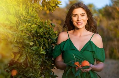 Kameraya bakarak ve gülümseyerek, bir turuncu meyve tutan genç kadın, açık havada bir portakal bahçesi nde günbatımı. Sağlıklı yaşam konsepti, cilt ve saç bakım kavramı.