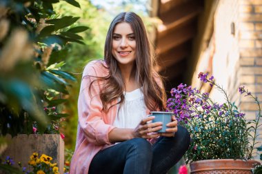 Gülümseyen ve kamera, ev terasta oturan bakarak güzel genç kız sıcak bir içecek içme bir kupa holding. Hobi ve istirahat eğlence kavramı.