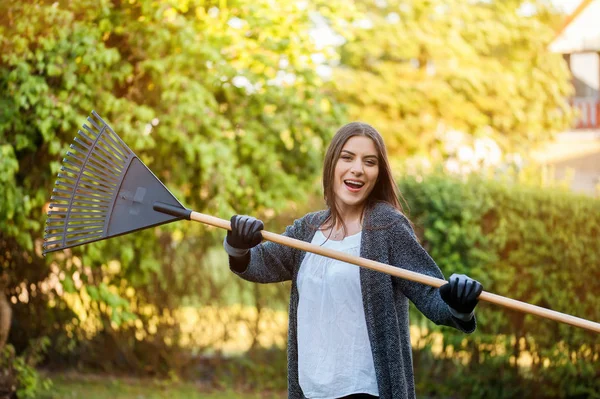 Smiling young woman with garden leaf rake in her home backyard with flowers, plants and vegetation. Gardening as hobby and leisure concept.