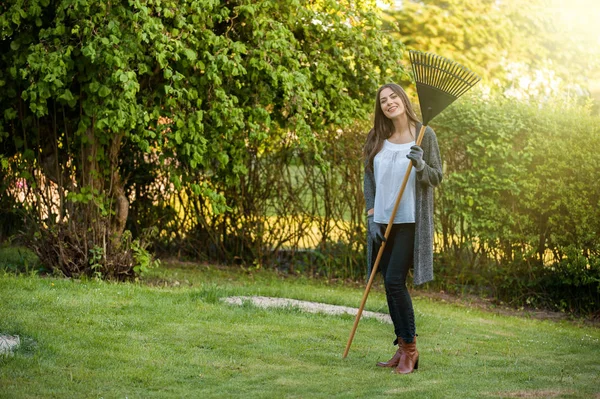 Smiling young woman with garden leaf rake in her home backyard with flowers, plants and vegetation. Gardening as hobby and leisure concept.