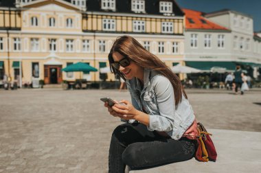 Bacaklar ile şehir merkezinde sokakta oturan güzel genç kadın güneşli gün ve cep telefonu tutarak ve gülümseyen üzerinde çarpı işareti.