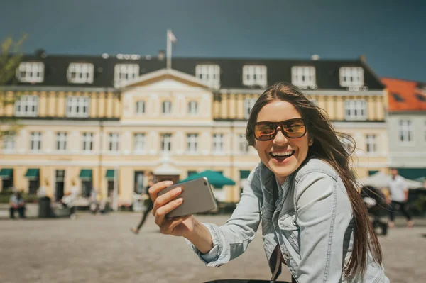 Young beautiful woman sitting in the city center taking a self portrait with her phone, smiling.
