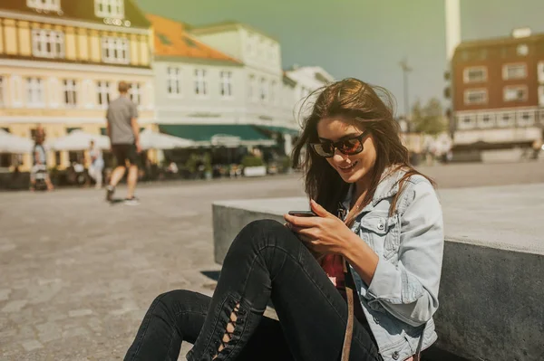 美丽的年轻妇女坐在城市中心的街道上一个晴朗的一天 手持手机和微笑 — 图库照片