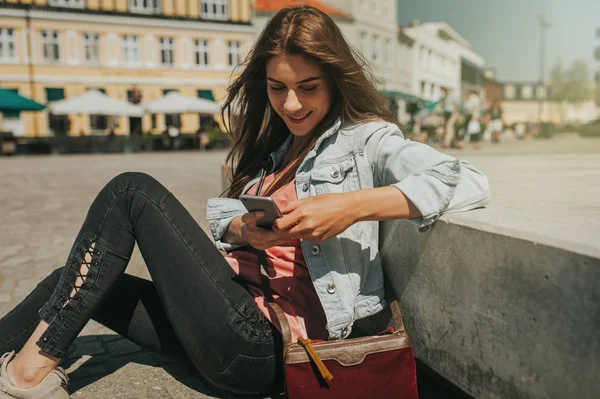 Beautiful young woman sitting on the street in city center with legs crossed on sunny day and holding mobile phone and smiling.