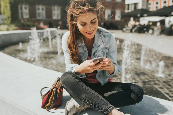 年轻的美女坐在市中心检查她的手机 — 图库照片