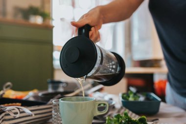 Kahvaltı masasının yakın çekimi ve elinde büyük bir kahve filtresi, gözenekli kahve fincanı olan bir kadın eli..