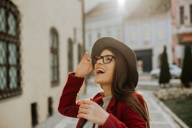 Şehirde mutlu genç hipster kadın gülümseyerek ve iyi vakit geçiriyor, şapkasını tutarak.