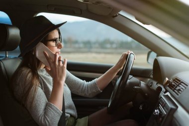 Gözlük ve şapka ile genç hipster kadın araba sürüş ve akıllı telefon tutarak