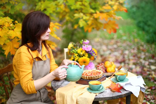 Autumn picnic. Woman in yellow dress and linen apron drinks tea from cup at wooden table in garden. Beautiful kettle, tablecloth, honey with spoon, apple pie, harvest, persimmon, grapes, maple leaf.