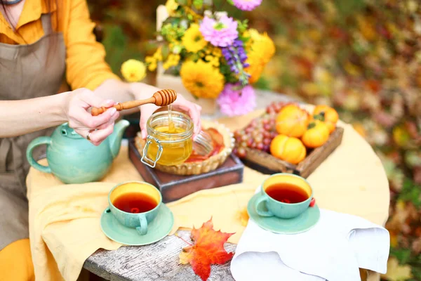 Autumn picnic. Woman in yellow dress and linen apron drinks tea from cup at wooden table in garden. Beautiful kettle, tablecloth, honey with spoon, apple pie, harvest, persimmon, grapes, maple leaf.