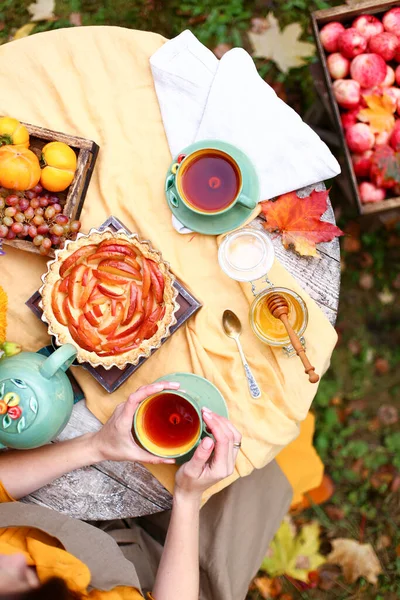 Autumn picnic. Woman in yellow dress and linen apron drinks tea from cup at wooden table in garden. Beautiful kettle, tablecloth, honey with spoon, apple pie, harvest, persimmon, grapes, maple leaf.