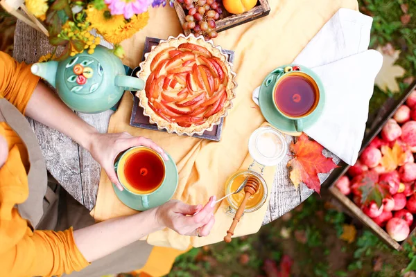 Autumn picnic. Woman in yellow dress and linen apron drinks tea from cup at wooden table in garden. Beautiful kettle, tablecloth, honey with spoon, apple pie, harvest, persimmon, grapes, maple leaf.