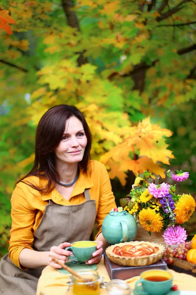 Autumn picnic. Woman in yellow dress and linen apron drinks tea from cup at wooden table in garden. Beautiful kettle, tablecloth, honey with spoon, apple pie, harvest, persimmon, grapes, maple leaf.