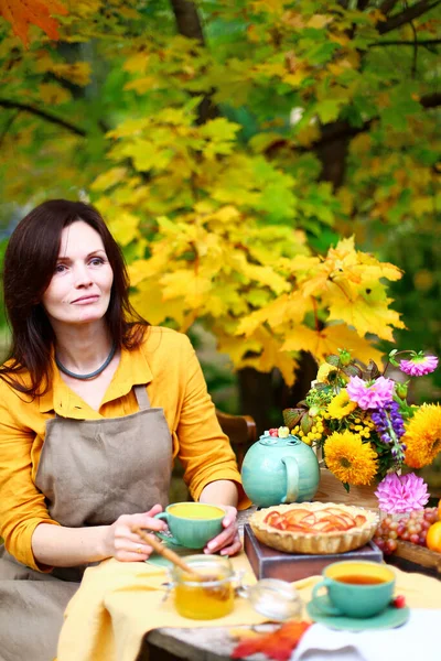 Autumn picnic. Woman in yellow dress and linen apron drinks tea from cup at wooden table in garden. Beautiful kettle, tablecloth, honey with spoon, apple pie, harvest, persimmon, grapes, maple leaf.