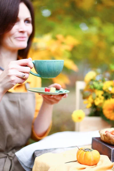 Autumn picnic. Woman in yellow dress and linen apron drinks tea from cup at wooden table in garden. Beautiful kettle, tablecloth, honey with spoon, apple pie, harvest, persimmon, grapes, maple leaf.