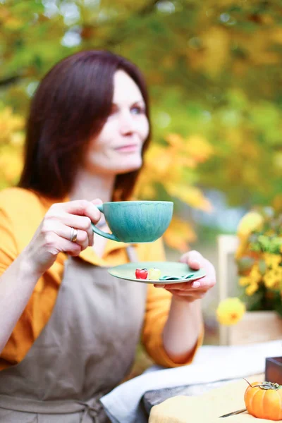 Autumn picnic. Woman in yellow dress and linen apron drinks tea from cup at wooden table in garden. Beautiful kettle, tablecloth, honey with spoon, apple pie, harvest, persimmon, grapes, maple leaf.