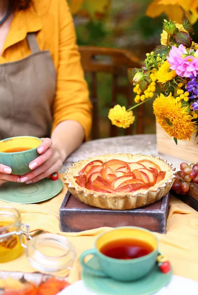 Autumn picnic. Woman in yellow dress and linen apron drinks tea from cup at wooden table in garden. Beautiful kettle, tablecloth, honey with spoon, apple pie, harvest, persimmon, grapes, maple leaf.