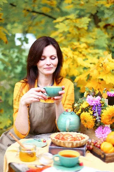 Autumn picnic. Woman in yellow dress and linen apron drinks tea from cup at wooden table in garden. Beautiful kettle, tablecloth, honey with spoon, apple pie, harvest, persimmon, grapes, maple leaf.