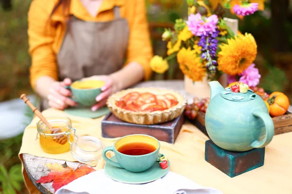 Autumn picnic. Woman in yellow dress and linen apron drinks tea from cup at wooden table in garden. Beautiful kettle, tablecloth, honey with spoon, apple pie, harvest, persimmon, grapes, maple leaf.