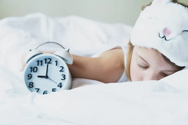 Bedtime. Little girl goes to sleep early evening with alarm clock on 9 p.m., wearing white cat, bunny sleep mask. Soft bed with white linen.Child sleep regime hours,daily routine.Sweet dreams concept.