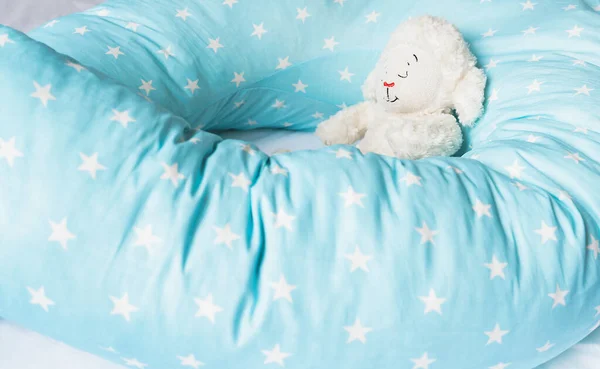 Light blue supporting pillow with stars on white sheet in bed for comfortable sleep of pregnant woman. Funny sleeping toy sheep, lamb. Goods, accessories for expectant mothers, kids, baby.