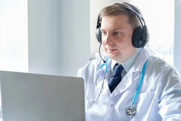 Doctor in headset speaks, talks to patient. Telehealth, telemedicine, online consultation, video call conference. Medical concept. Caucasian man,blue eyes,white robe,stethoscope. Coronavirus covid-19.