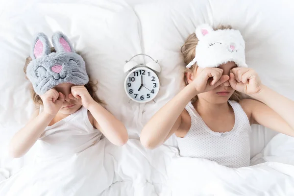 Little girl, sisters sleep in bed with big alarm, clock, rub eyes with fists. Soft cat, bunny mask. Early morning rise to kindergarten, study to school. Evening bedtime. Kids daily routine for child.
