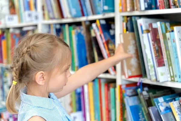 Little girl chooses, takes book with fairy tales from shelf in children\'s library.Special reading kids room.Many shelves with bright interesting,educational books about animals.
