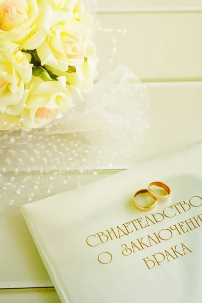 Golden wedding rings,marriage certificate and bride bouquet on a table of light wood. Indoors. Vertical format. Color. Photo.