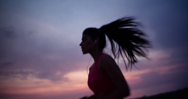 Νεαρή δρομέας γυναίκα με ακουστικά τρέχει στο ηλιοβασίλεμα — Αρχείο Βίντεο