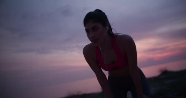 Νεαρή γυναίκα σε φόρμα με ακουστικά που ξεκουράζονται μετά το τρέξιμο στο ηλιοβασίλεμα — Αρχείο Βίντεο