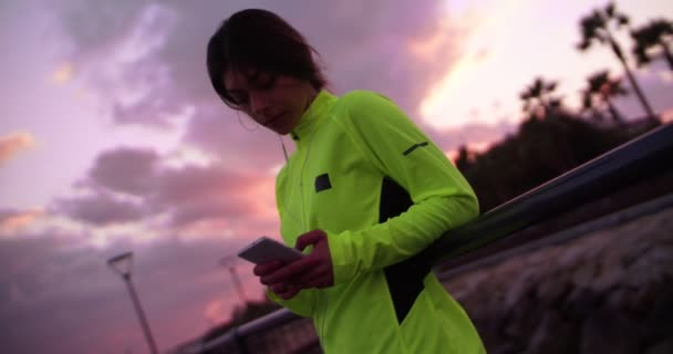 Mujer joven en ropa deportiva mensajes de texto en su teléfono al atardecer — Vídeos de Stock