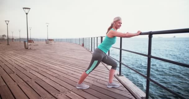 Actieve senior vrouw met koptelefoon stretching voor joggen aan zee — Stockvideo