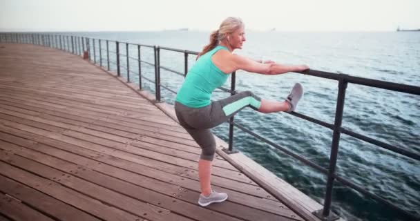 Sportliche Seniorin in Sportbekleidung streckt ihre Beine auf Pier — Stockvideo