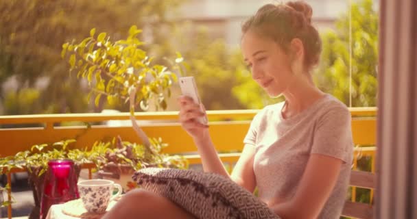 Joven mujer hipster sentado en el balcón y mensajes de texto en el teléfono inteligente — Vídeos de Stock