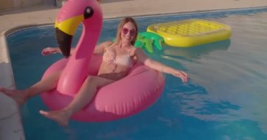 Yüzme havuzunda şişme pembe flamingoda yüzen genç bir kadın.