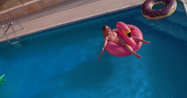 Ung kvinne som flyter på rosa oppblåsbar flamingo i svømmebasseng – stockvideo