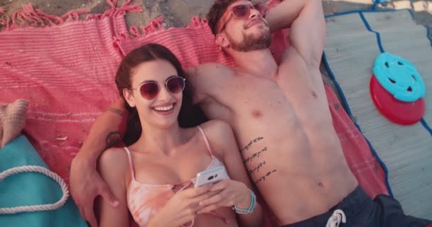 Joven pareja hipster relajarse tumbado en la playa y mensajes de texto — Vídeo de stock