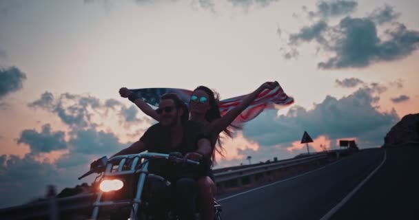 Joven pareja de motociclistas montando en moto y ondeando bandera estadounidense — Vídeo de stock