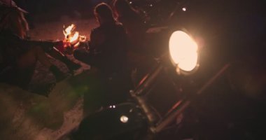 Motosikletli asi arkadaşlar şenlik ateşi etrafında oturup içiyorlar.