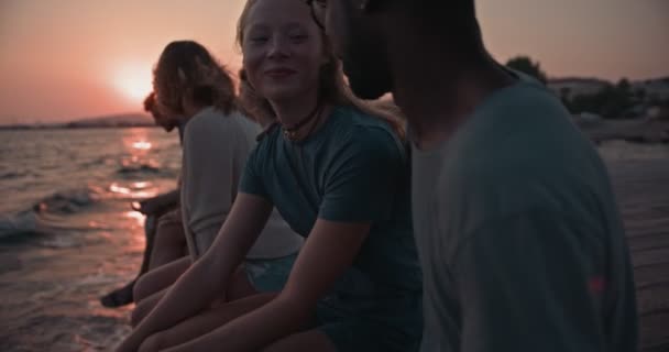 Młodzi, wieloetniczni przyjaciele i pary siedzące na molo o zachodzie słońca — Wideo stockowe