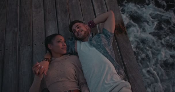 Fiatal romantikus, többnemzetiségű pár pihenés a fa mólón naplementekor — Stock videók