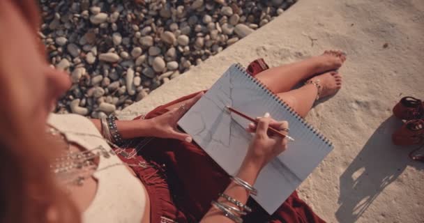 Νεαρή μποέμικη γυναίκα ζωγραφίζει σε μπλοκ ζωγραφικής στην παραλία — Αρχείο Βίντεο
