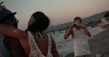 Çok ırklı arkadaşlar plajda klasik kamerayla tatil fotoğrafları çekiyorlar.