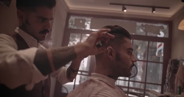 Молодий стильний старомодний перукар з татуюваннями, що ріжуть волосся клієнтів — стокове відео