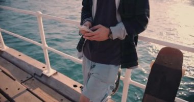 Genç hippi kaykaycı iskelede duruyor ve akıllı telefondan mesaj atıyor.