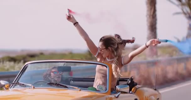 Mujeres jóvenes conduciendo un coche descapotable y celebrando con bombas de humo — Vídeo de stock