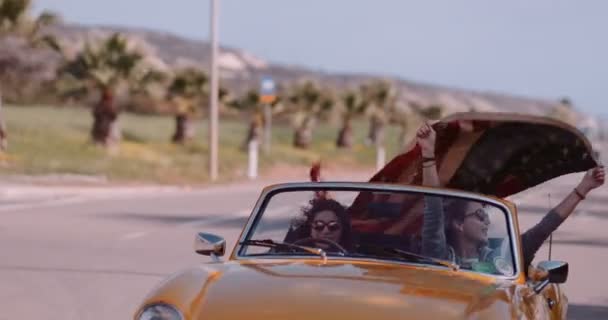 Mujeres jóvenes en amarillo retro convertible celebrando con bandera americana — Vídeo de stock