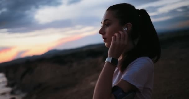 Jonge vrouwelijke loper ontspannen met muziek op het strand bij zonsondergang — Stockvideo
