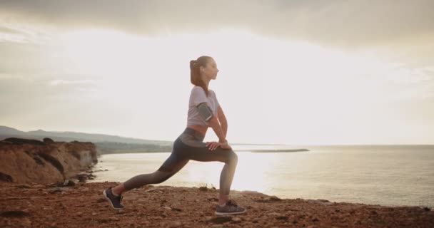 Sportovní žena protahuje nohy na mořském útesu při západu slunce — Stock video